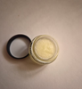 An open clear cosmetic jar filled with a pale yellow balm or scrub, shown with its black lid placed beside it on a plain beige surface.