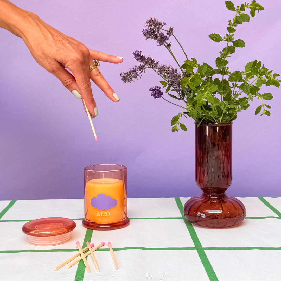 Hand lighting a candle with a purple logo, next to a vase with flowers on a white and green checkered surface.