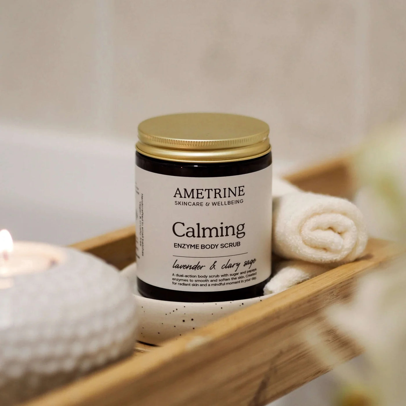 Ametrine Calming Enzyme Body Scrub jar on a wooden tray with towels and a candle.