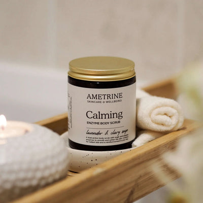 Ametrine Calming Enzyme Body Scrub jar on a wooden tray with towels and a candle.
