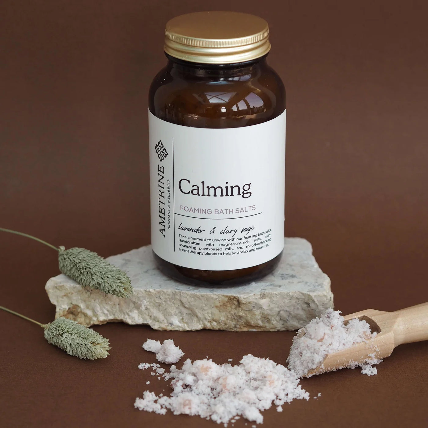 Bottle of 'Calming' foaming bath salts with a wooden scoop and herbs on a brown background