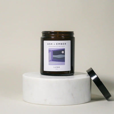 Candle jar labeled 'ASH + Ember' on a white pedestal with a neutral background