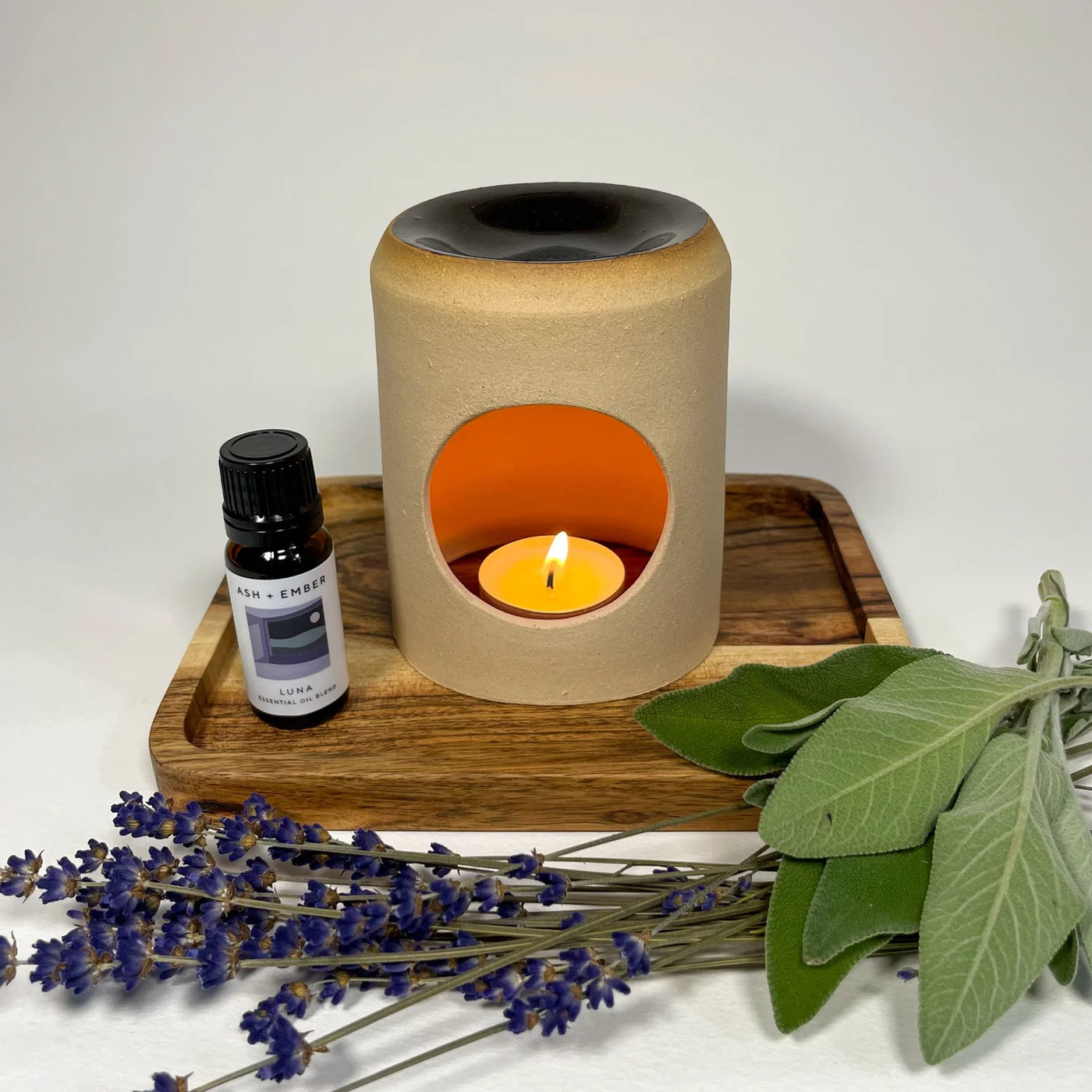 Aroma lamp with a lit candle on a wooden tray, surrounded by lavender and green leaves.