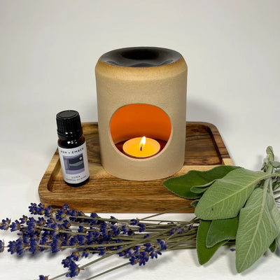 Aroma lamp with a lit candle on a wooden tray, surrounded by lavender and green leaves.