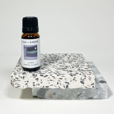 Essential oil bottle labeled 'Luna' by 'Ash + Ember' on a terrazzo-patterned stand.