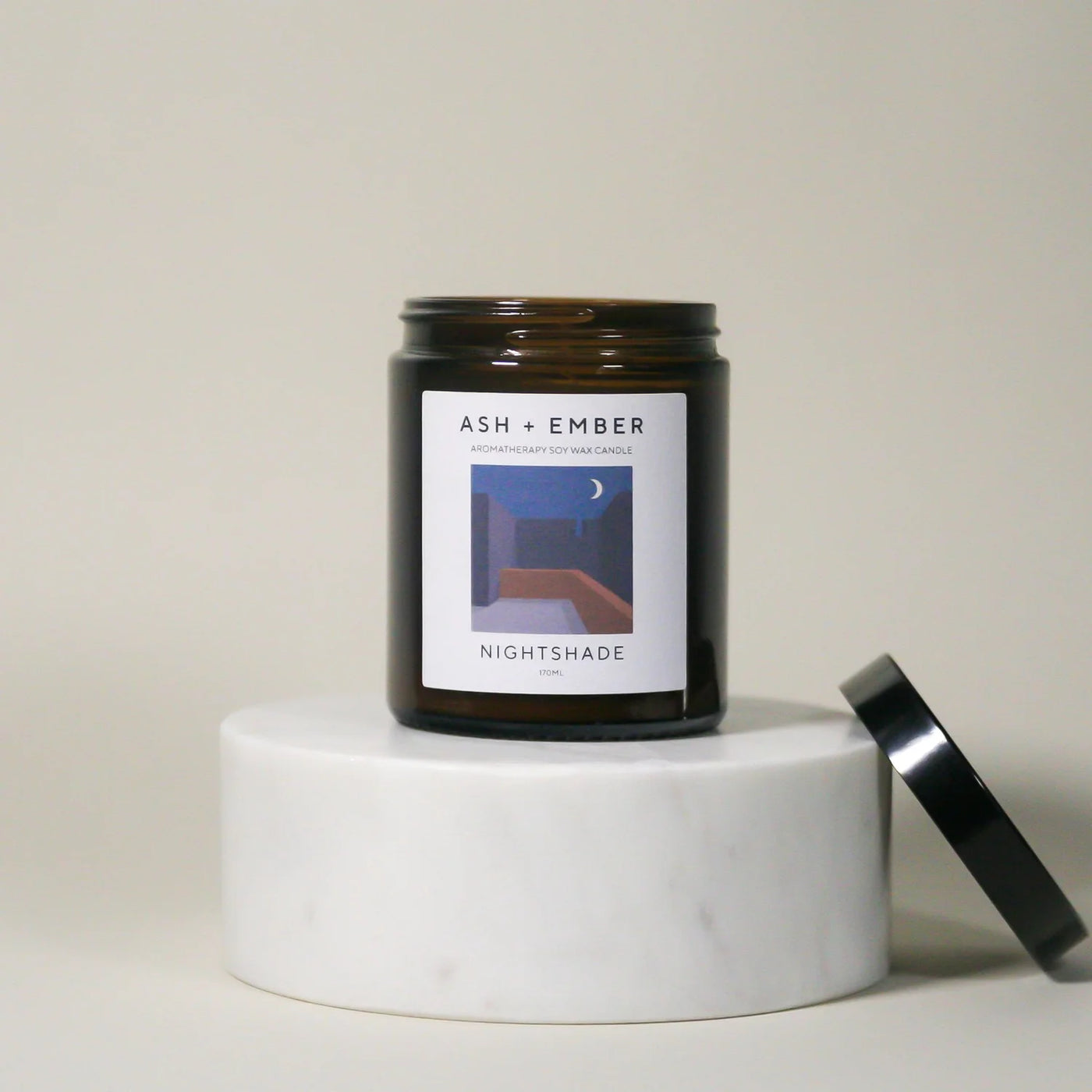 Candle jar labeled 'Ash + Ember Nightshade' on a white pedestal with a neutral background
