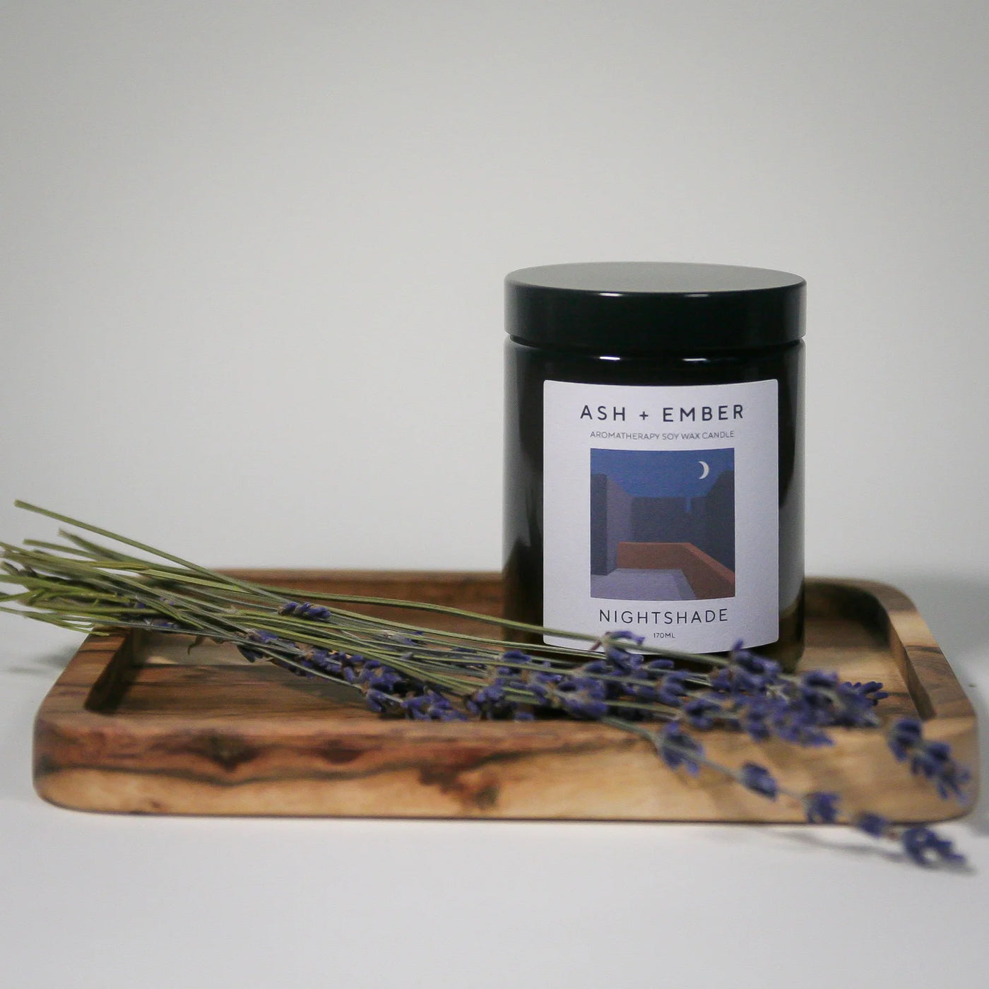 Candle jar labeled 'ASH + Ember Nightshade' on a wooden tray with lavender.