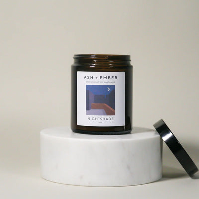 Candle jar labeled 'Ash + Ember Nightshade' on a white pedestal with a neutral background
