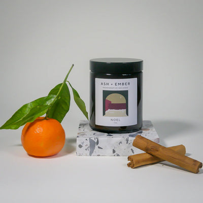 Candle labeled 'ASH + Ember' with an orange, cinnamon stick, and leaves on a white background