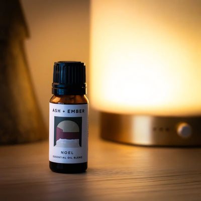 Essential oil blend bottle labeled 'ASH + EMBER NOEL' on a wooden surface with a warm light in the background.