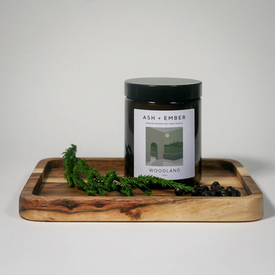 Candle jar labeled 'Woodland' on a wooden tray with greenery against a light background