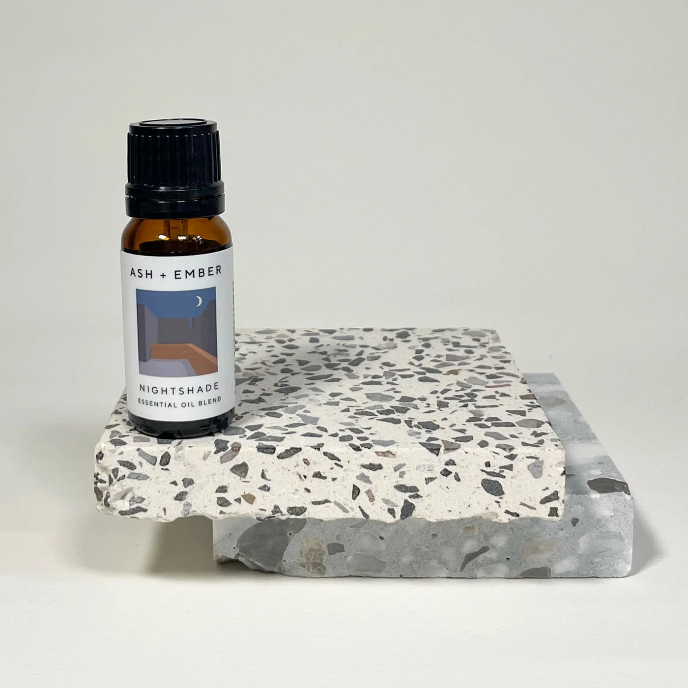 Essential oil bottle labeled 'Ash + Ember' on a terrazzo coaster with a white background