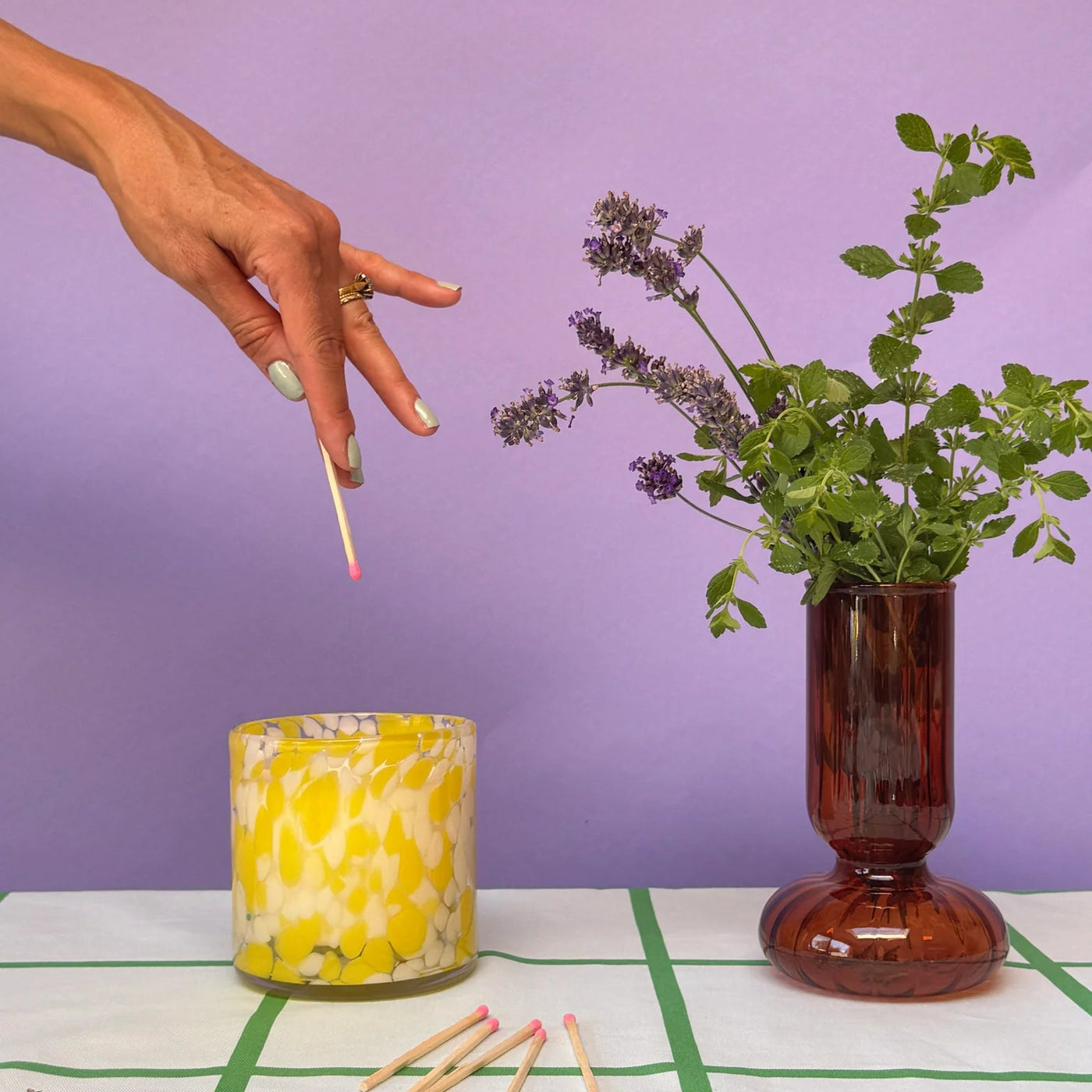 Hand lighting a candle next to a vase with greenery on a checkered tablecloth against a purple background