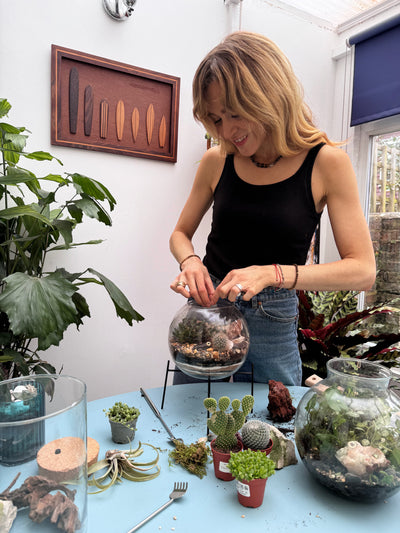 Person arranging plants in a terrarium on a table with various gardening tools and materials.