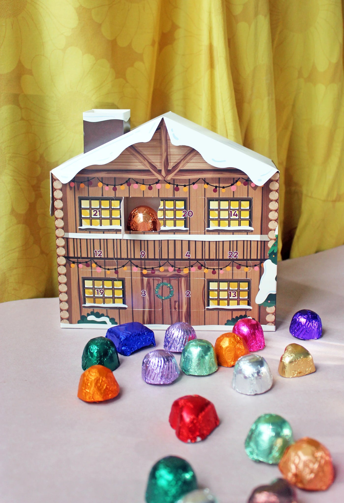 Wooden house-shaped Advent calendar with colorful chocolate eggs on a yellow curtain background