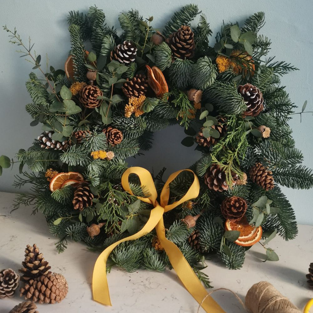 Christmas wreath with pine cones, oranges, and a yellow ribbon on a light surface.
