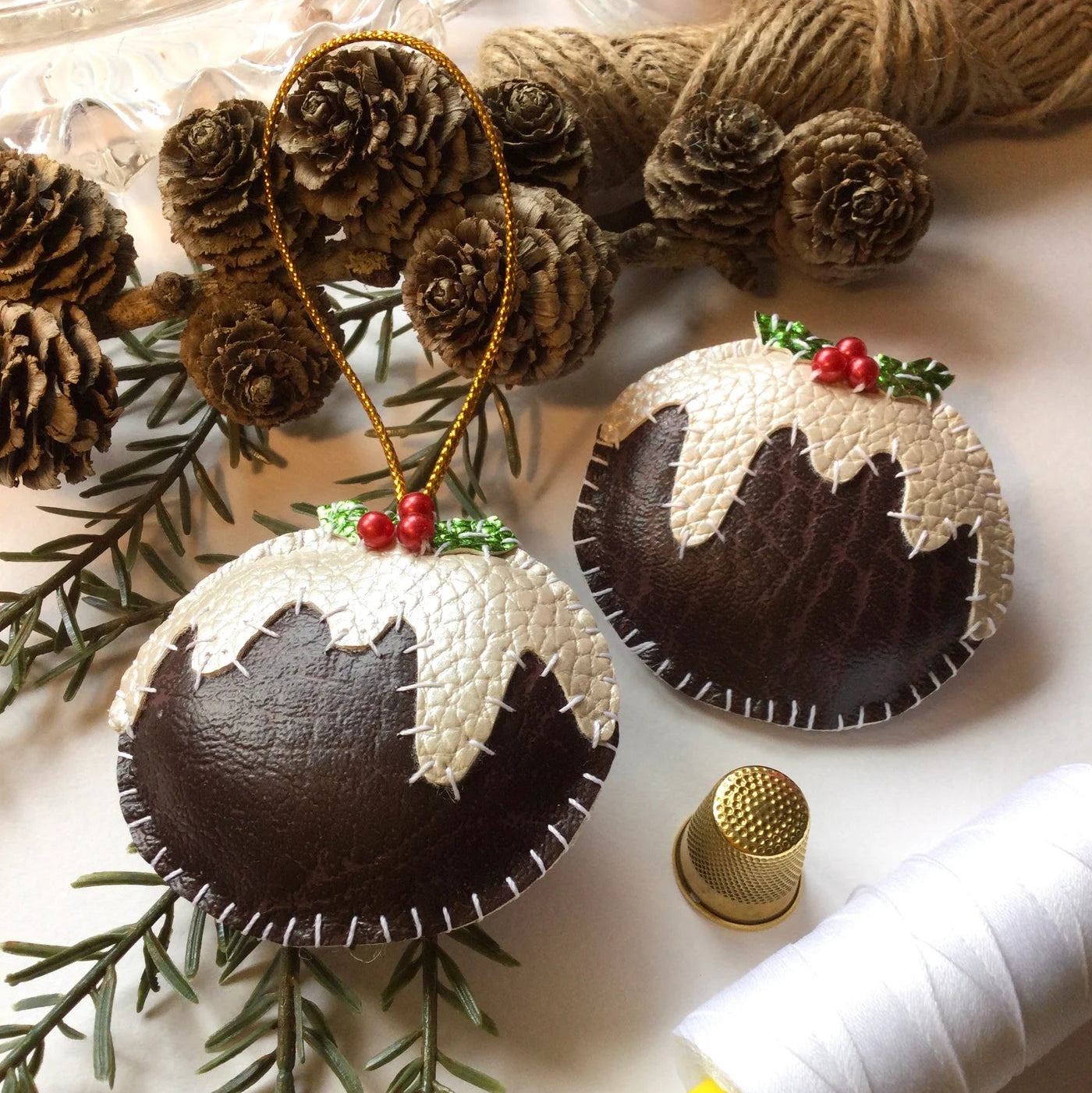 Christmas-themed fabric ornaments resembling Christmas puddings on a white surface with pine branches and thread spool.