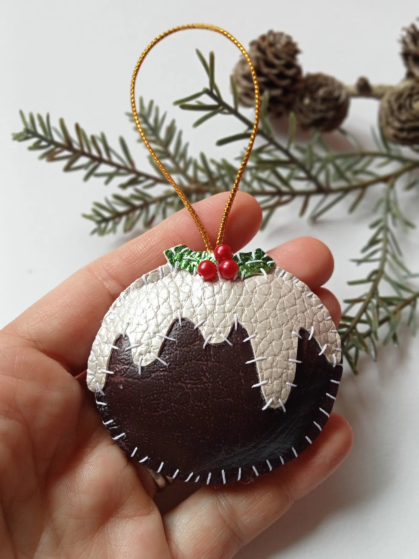 Hand holding a Christmas pudding ornament with gold string, green leaves, and red berries against a white background.