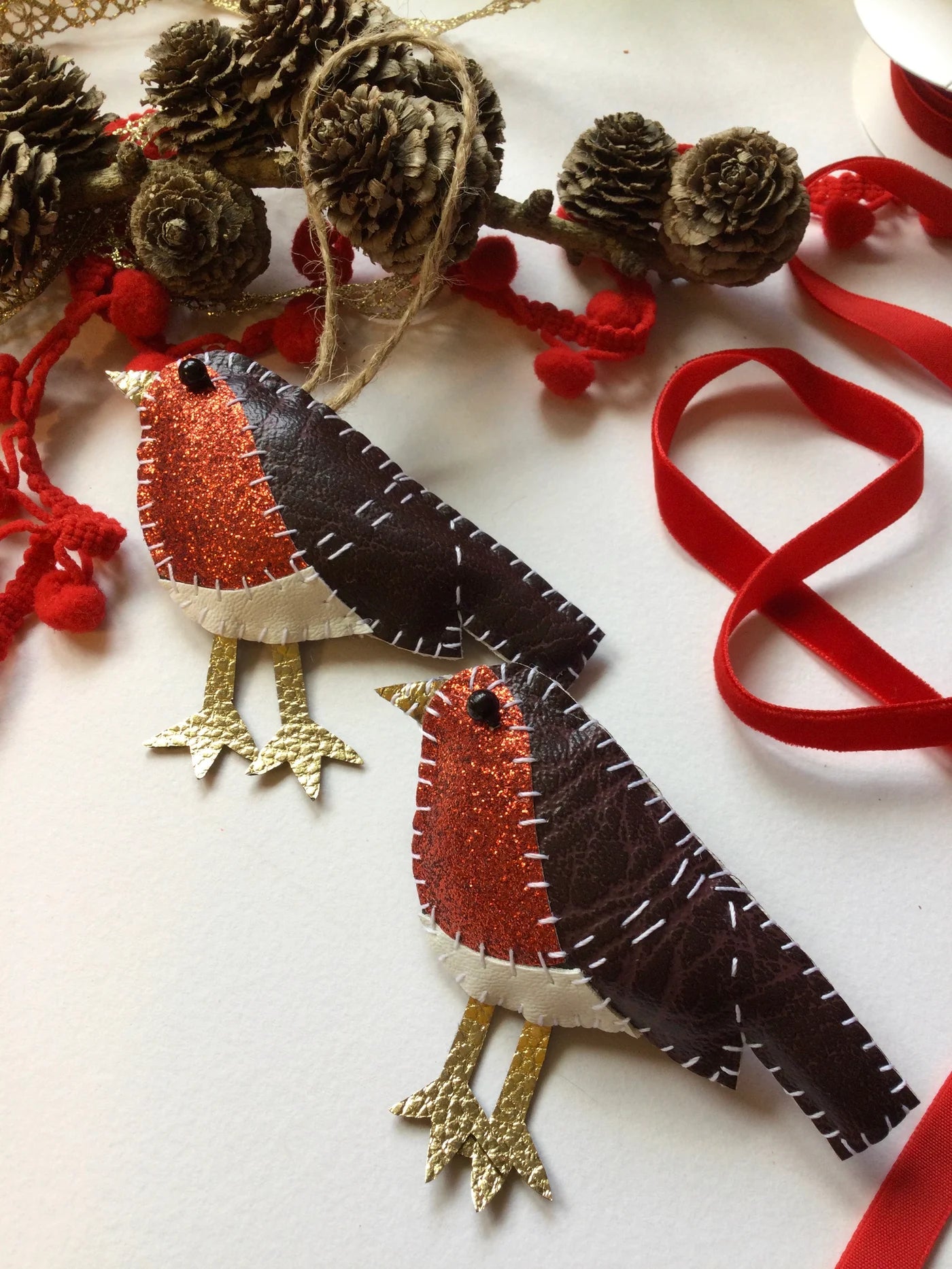 Two felt robin decorations on a white background with red ribbons and pine cones.