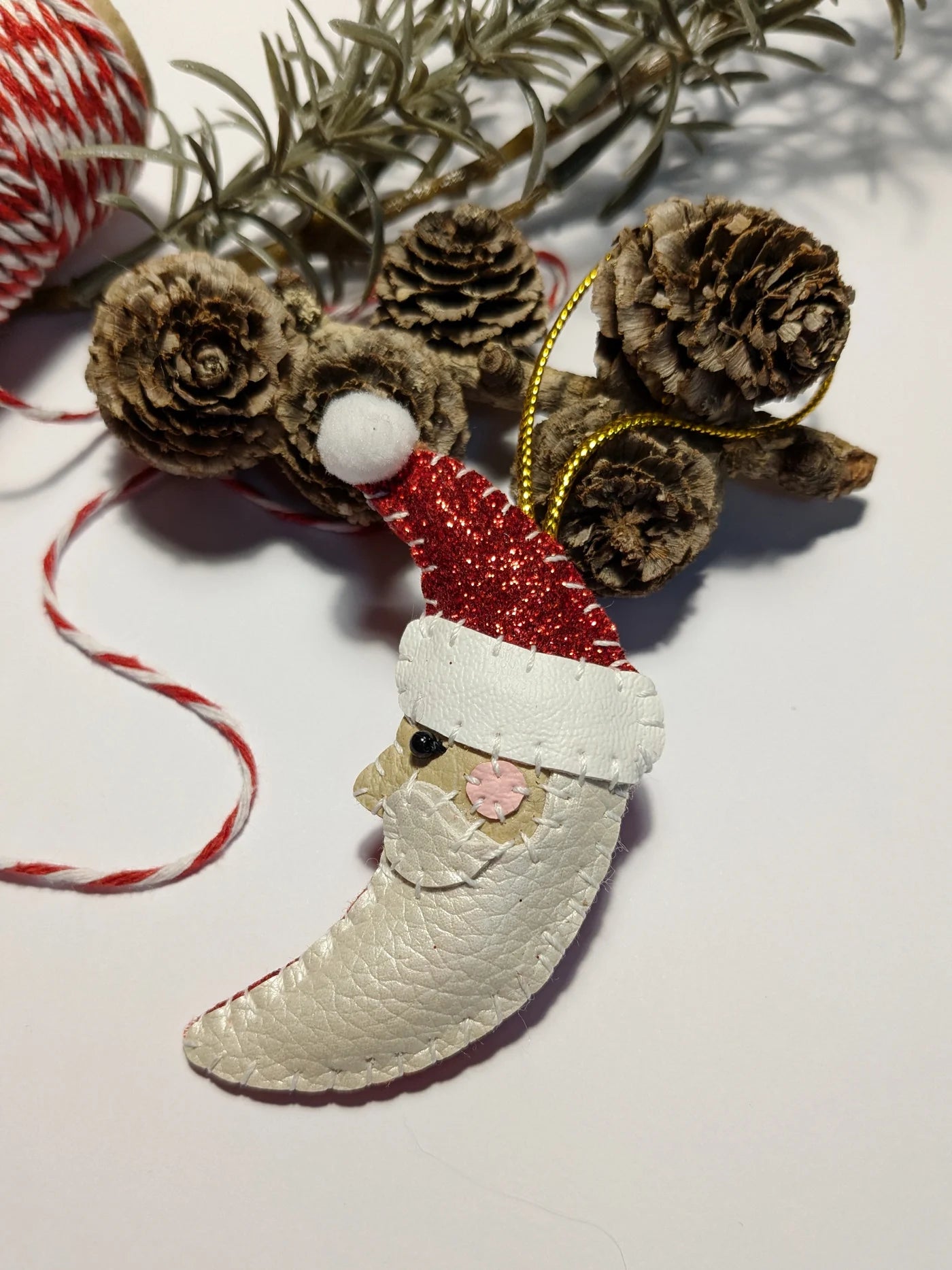 Decorative ornament shaped like a boot with a Santa hat, surrounded by pine cones and branches on a light background.