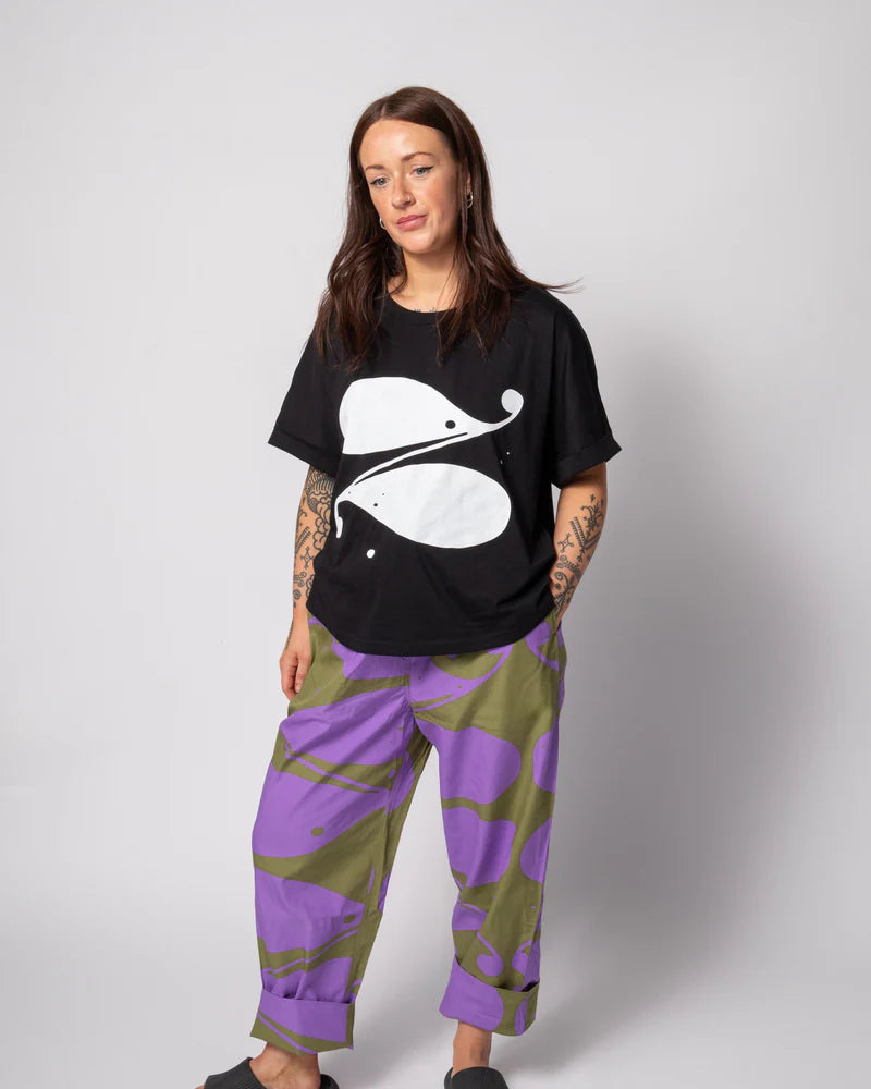 Person wearing a black t-shirt with a white graphic and purple and green pants on a plain background