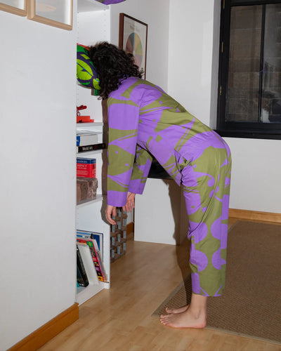 Person wearing a purple and green patterned outfit bending over in a room with a bookshelf.