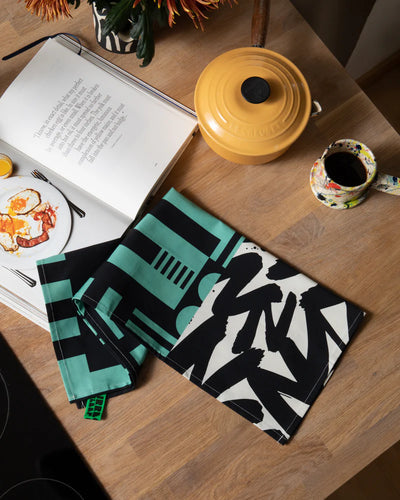 Two patterned tea towels on a wooden table with a book and coffee cup.