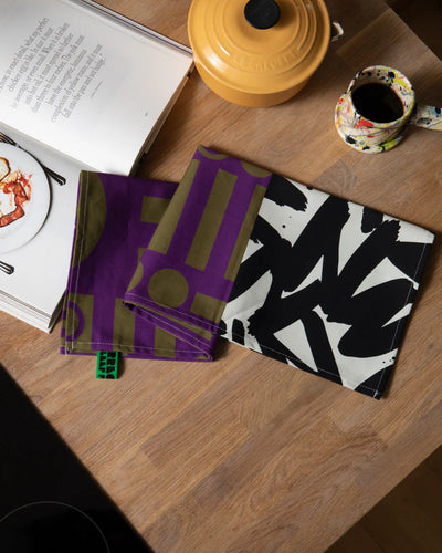 Two patterned tea towels on a wooden surface with a book and yellow container in the background.