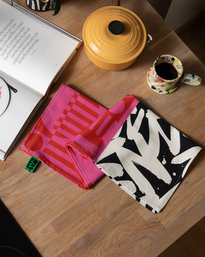 One patterned tea towel on a wooden surface with a book and yellow pot in the background.
