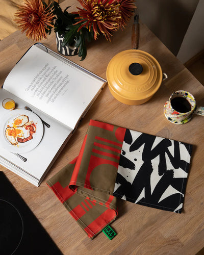 Tea towels with geometric patterns on a wooden table with a book and cup.