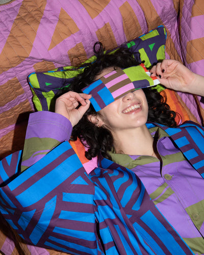 Person wearing a colorful eye mask with a pattern of geometric shapes, lying on a bed with a multicolored quilt.