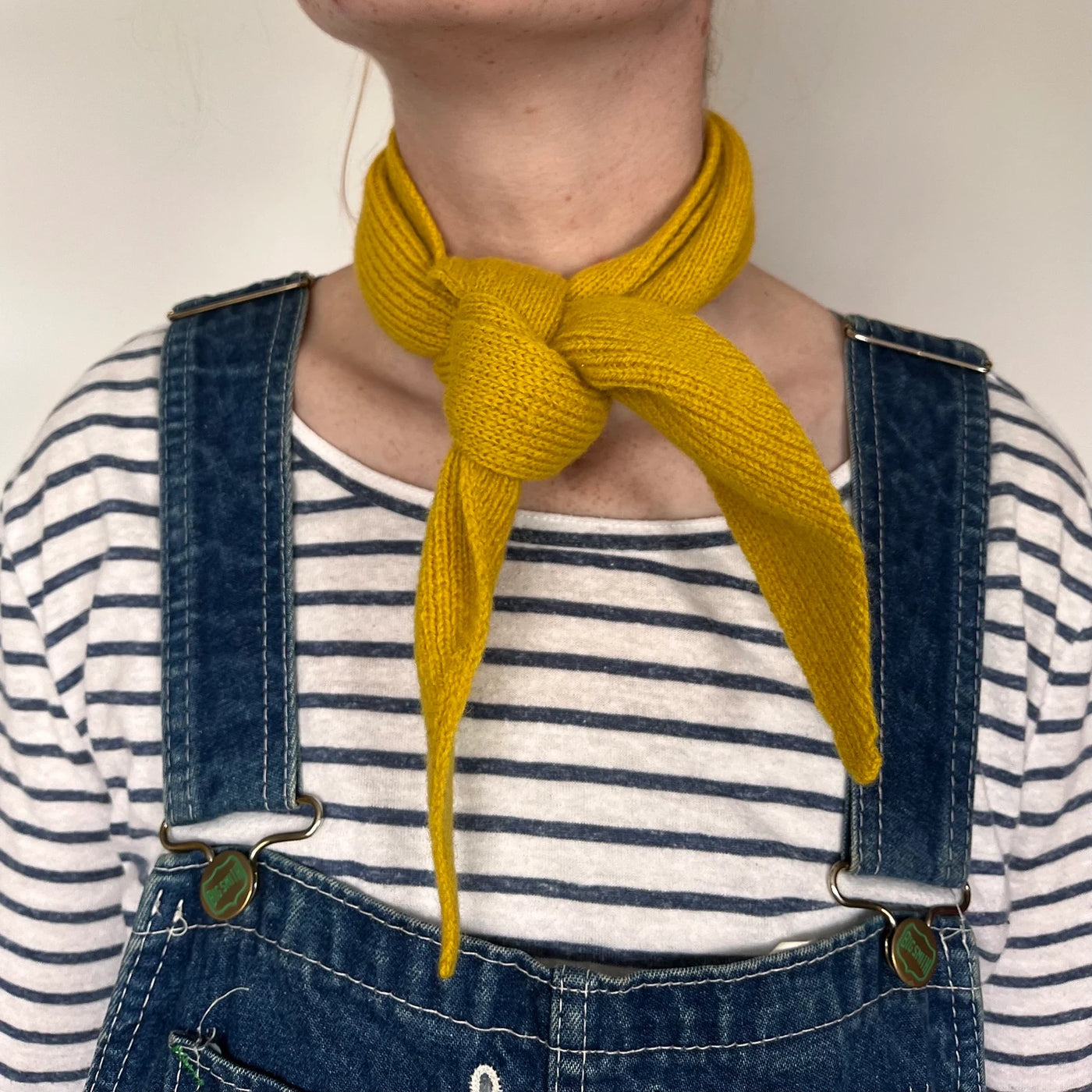 Person wearing a yellow knitted scarf, striped shirt, and denim overalls against a neutral background