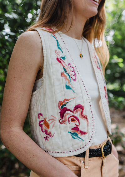 Woman wearing a white sleeveless top with colorful floral embroidery in an outdoor setting.