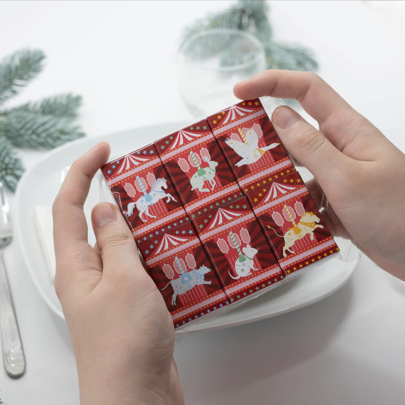 Hands holding a red patterned box with Christmas-themed illustrations on a white surface.