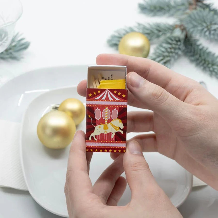 Hand holding a matchbox with a festive design, surrounded by gold ornaments and greenery on a white surface.