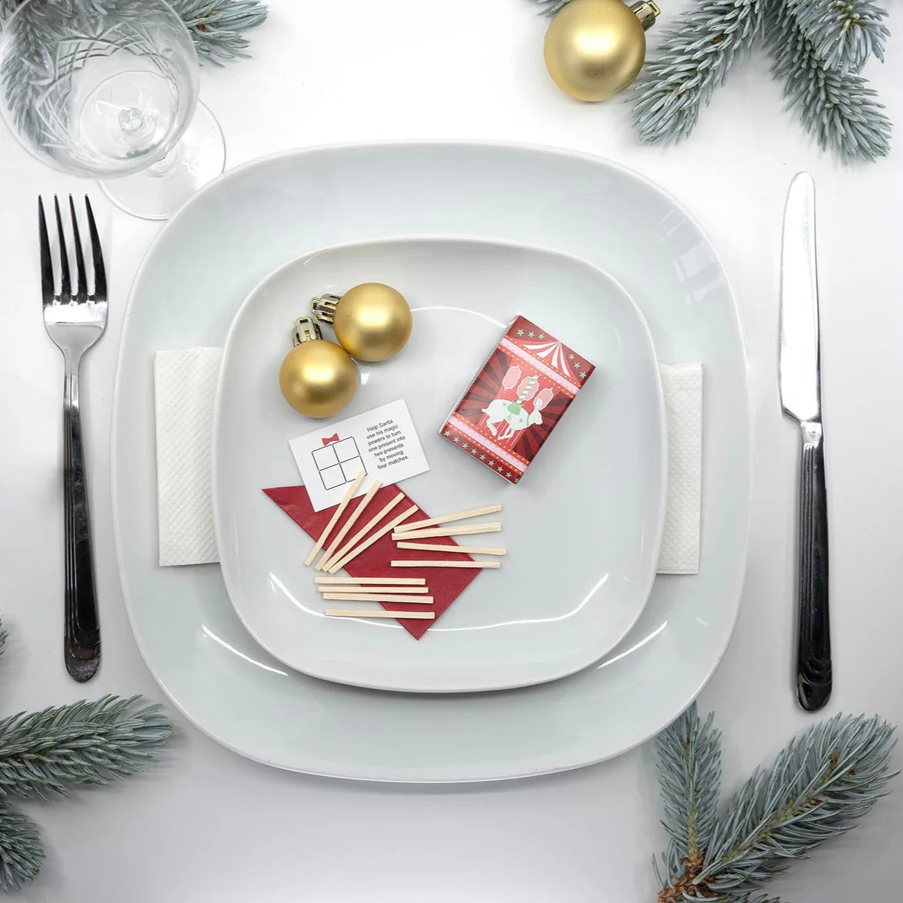 Christmas-themed table setting with gold ornaments, red cards, and greenery on a white surface.