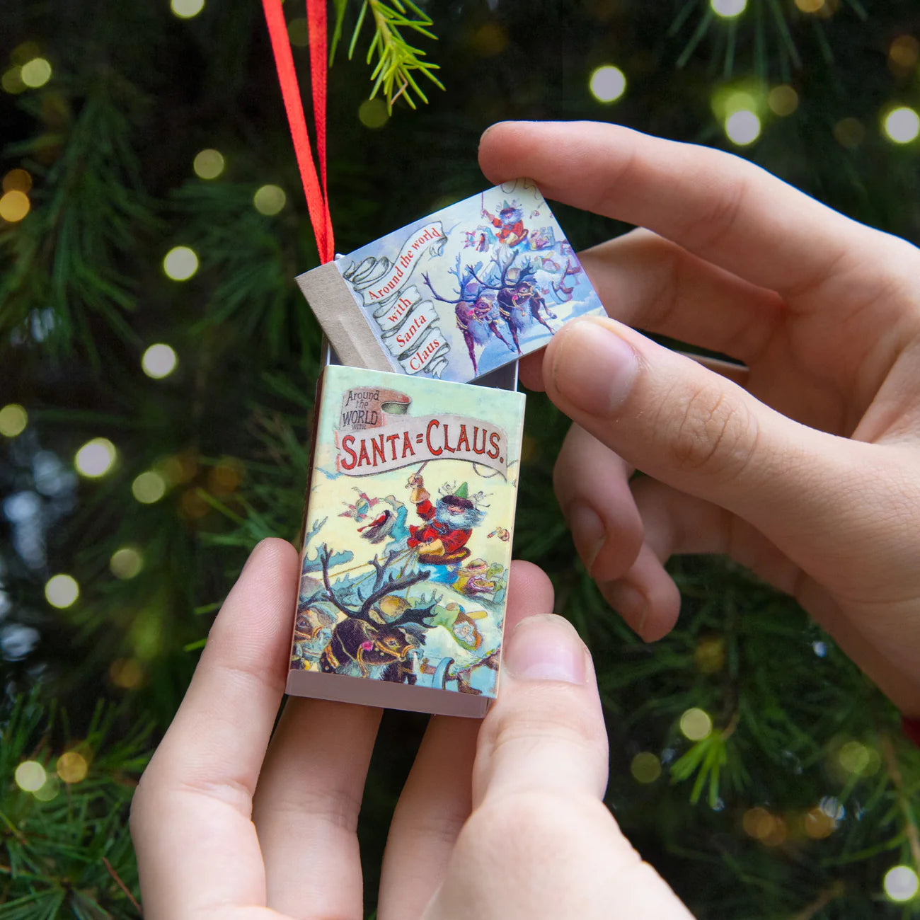 Miniature book ornament with 'Santa Claus' design held by hands against a Christmas tree background.