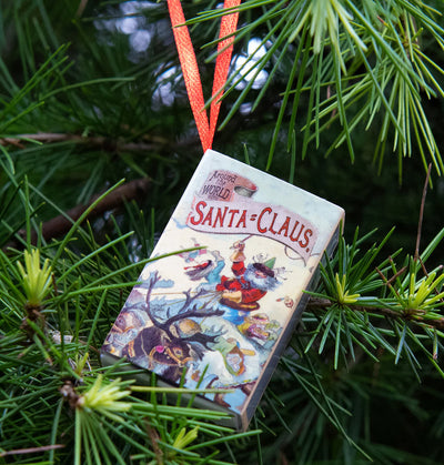 Book-shaped ornament with 'Santa-Claus' text and illustration, hanging on a Christmas tree.