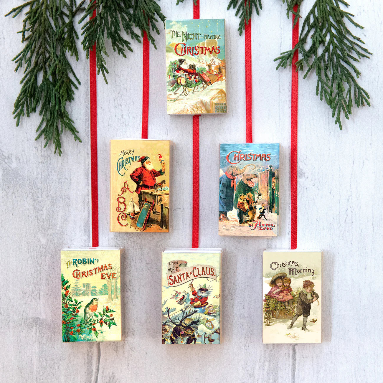 Vintage-style Christmas book covers hanging on a white wall with red ribbons and greenery.