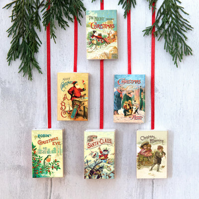 Vintage-style Christmas book covers hanging on a white wall with red ribbons and greenery.