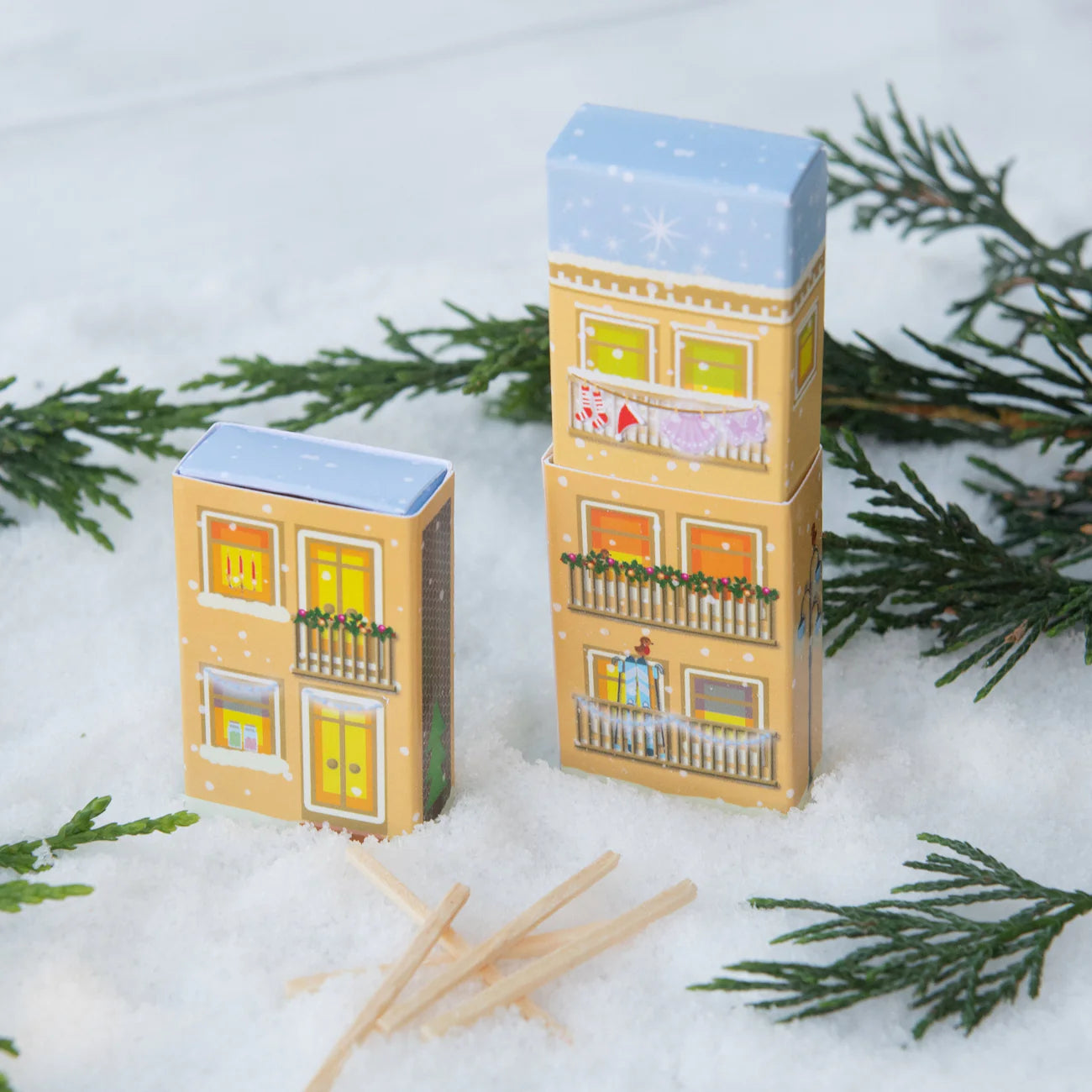 Two paper house models on a snowy surface with evergreen branches.
