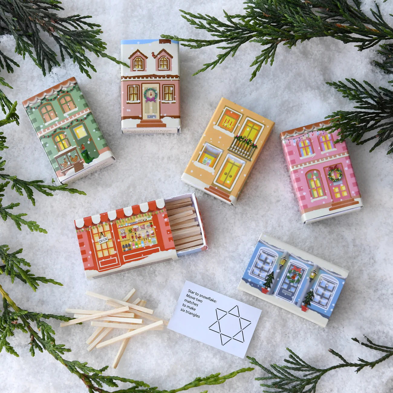 Matchboxes with house designs on a snowy surface with matchsticks and greenery.