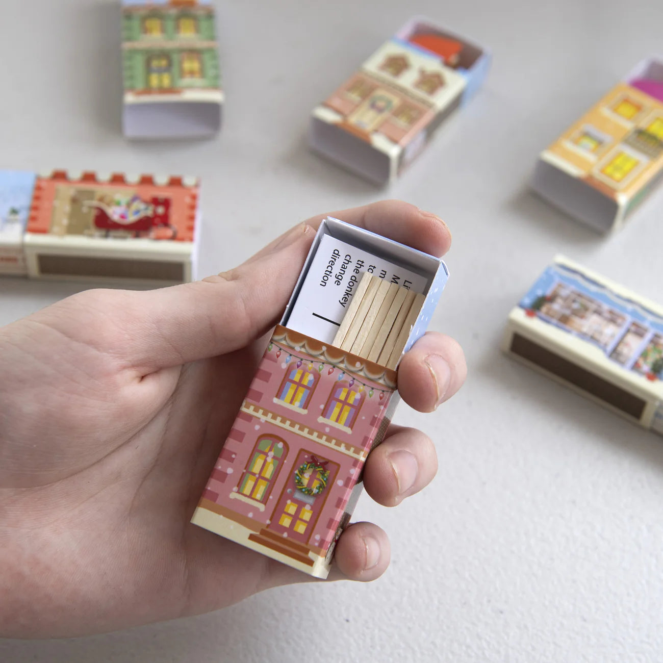 Hand holding a matchbox with decorative design, surrounded by more matchboxes on a light background