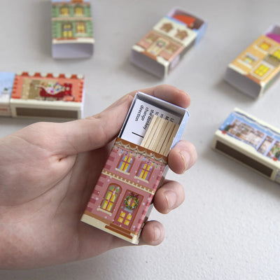 Hand holding a matchbox with decorative design, surrounded by more matchboxes on a light background
