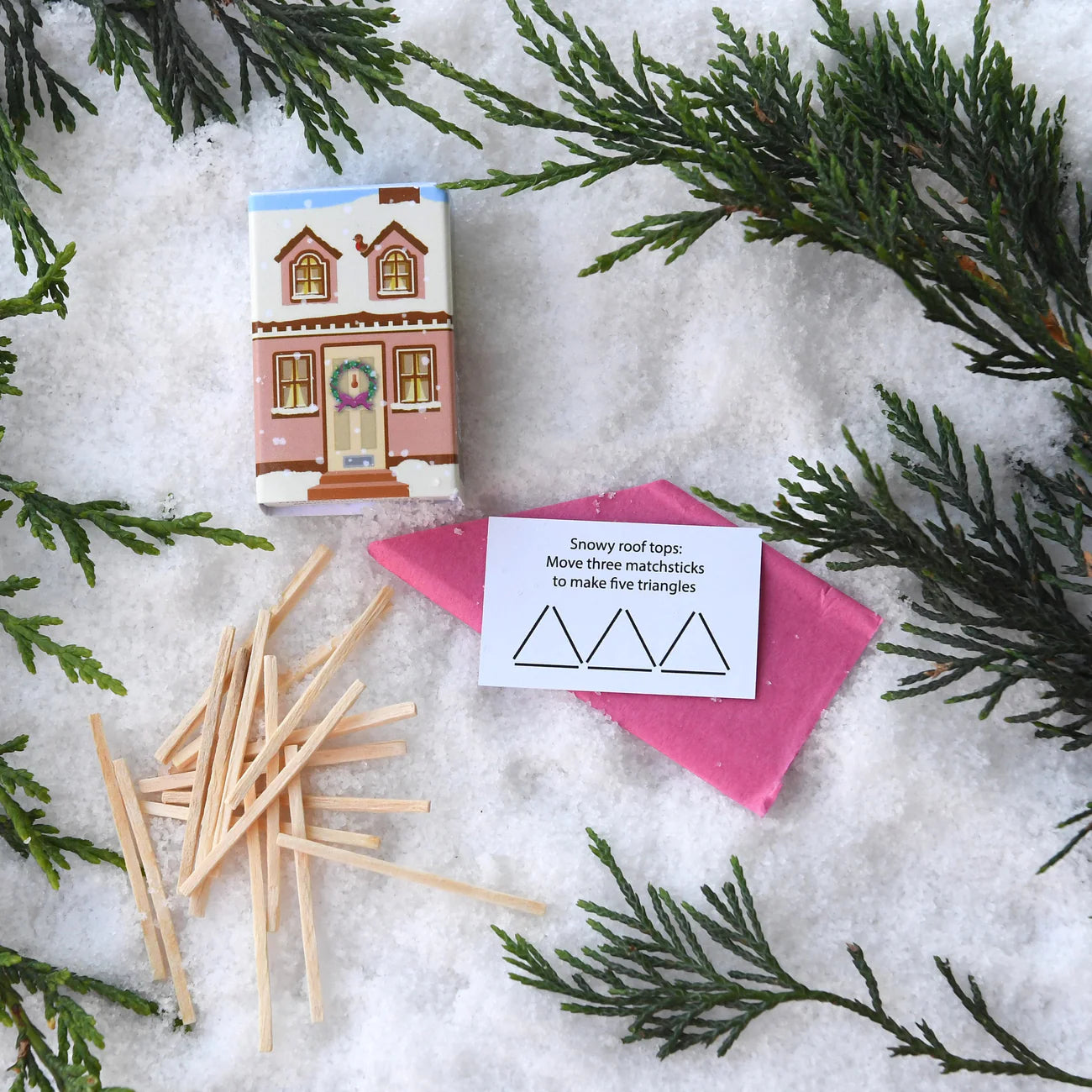 Matchstick house, matchsticks, and a card on a snowy background with evergreen branches.