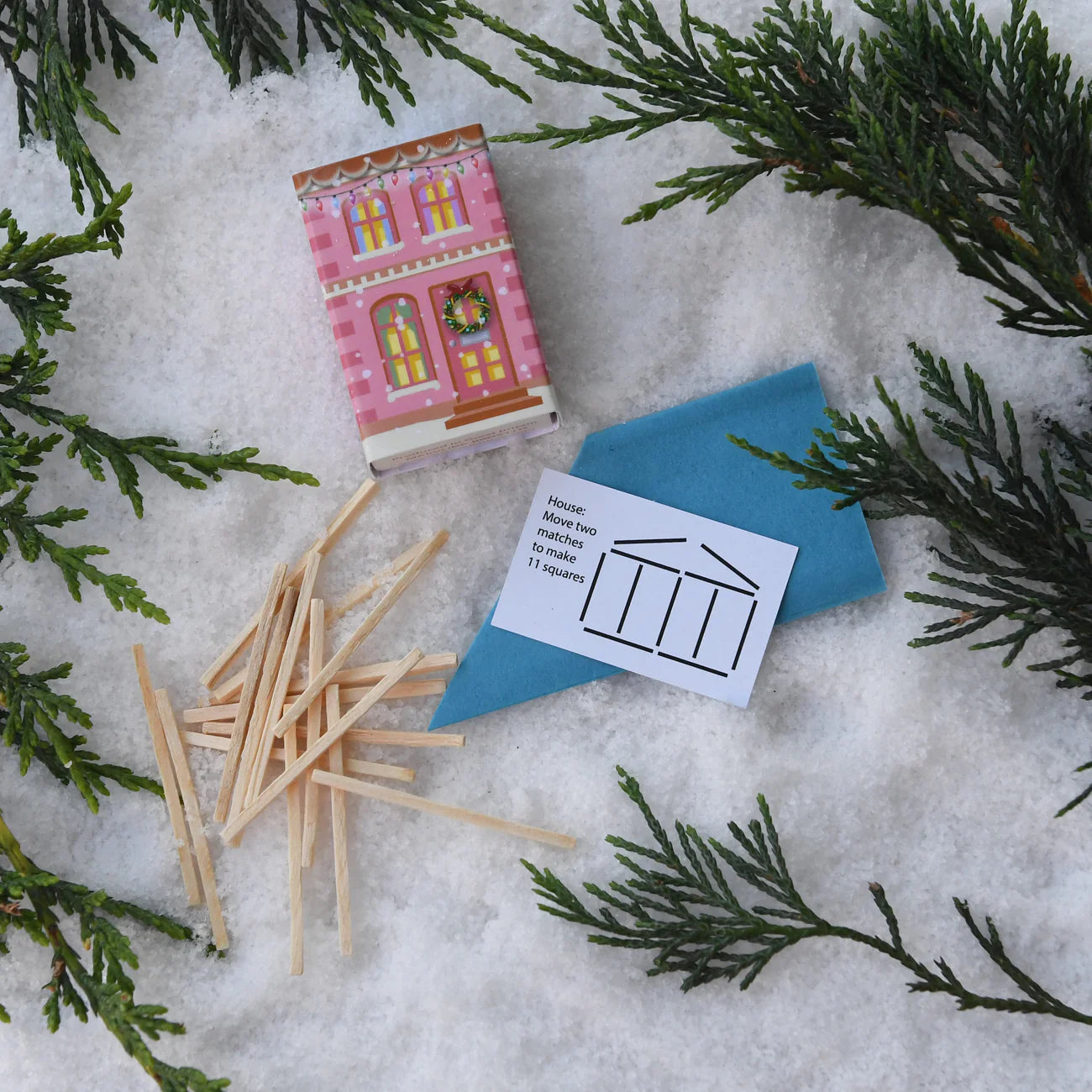 Matchbook with house design, matches, and blue envelope on snow with evergreen branches