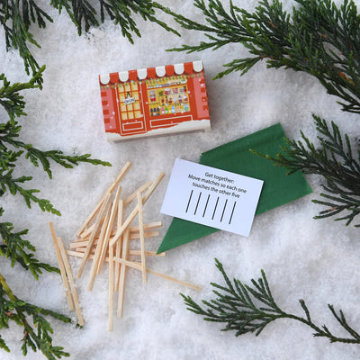 Box of matches, green paper, and wooden sticks on a snowy surface with evergreen branches.