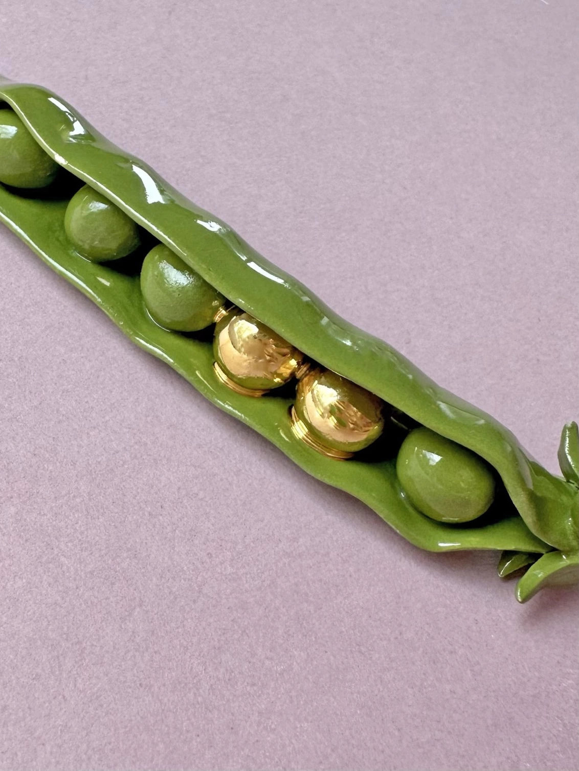 Brooch shaped like a green pea pod with gold peas on a light purple background