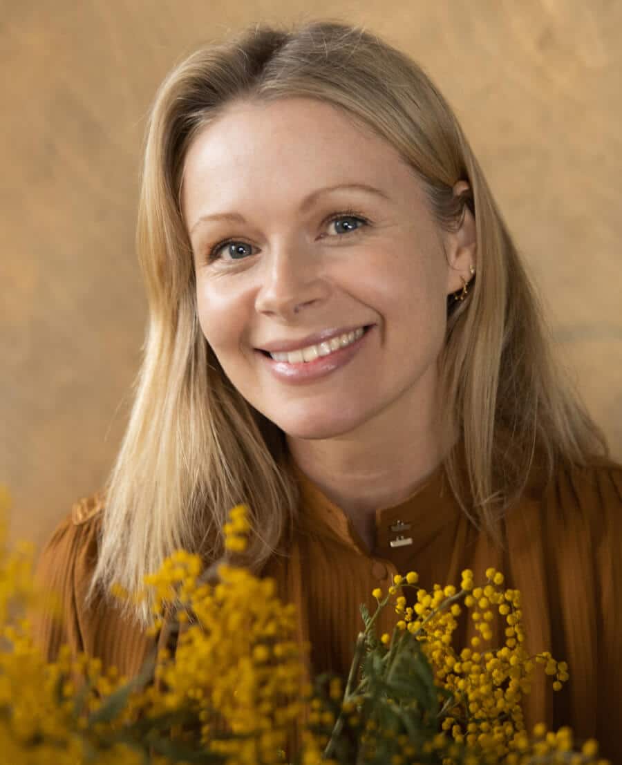 Woman with blonde hair and a brown top holding yellow flowers against a beige background