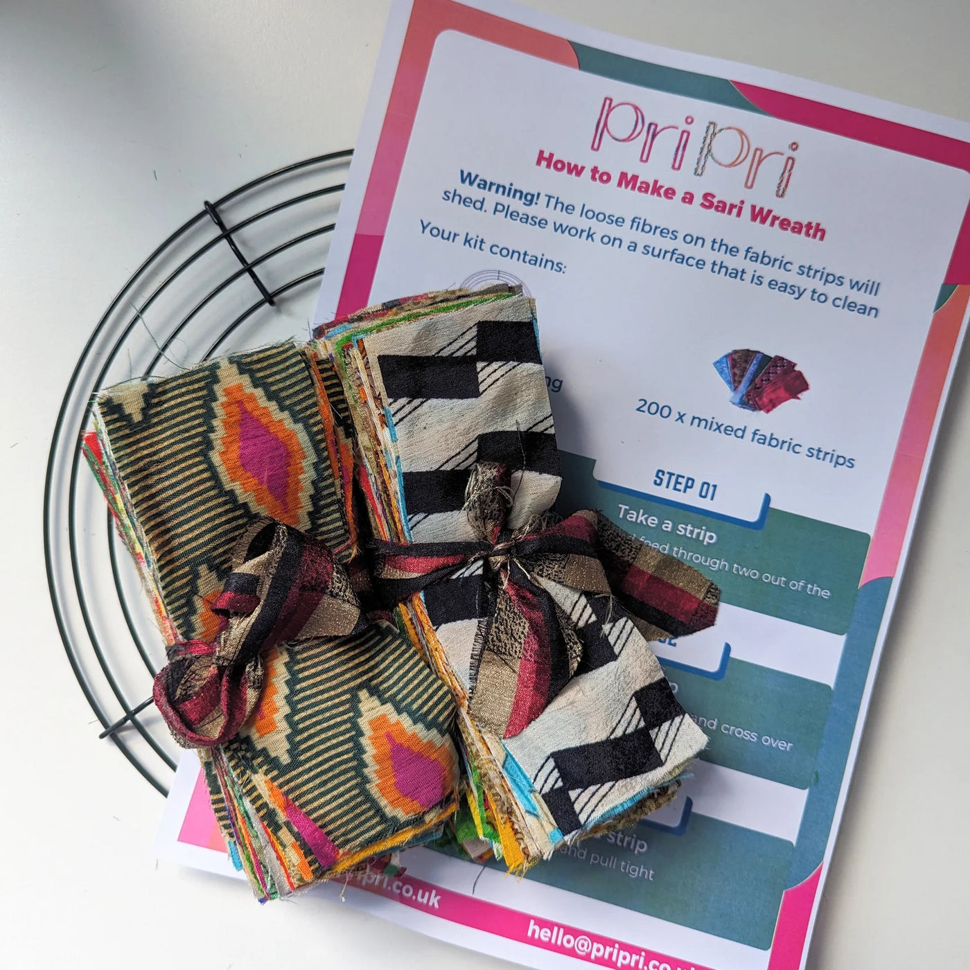 Colorful fabric strips and a 'priPri' kit for making a sari wreath on a white surface.